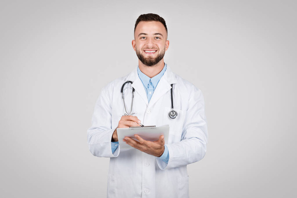 Vrolijke mannelijke arts met een klembord voor patiëntendossiers, met een stethoscoop om zijn nek, die professionaliteit in de gezondheidszorg vertegenwoordigt - Foto, afbeelding