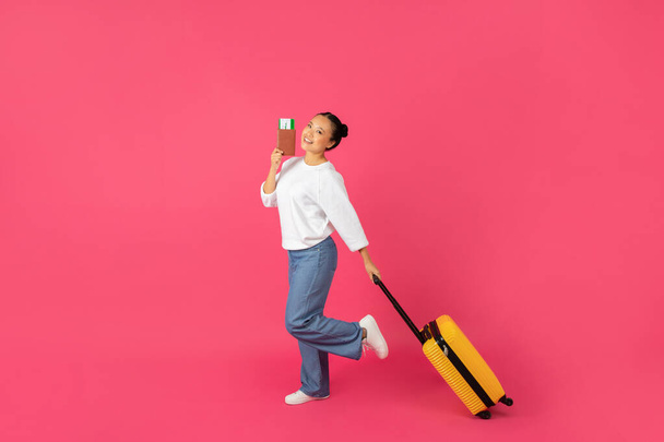 Koncepcja podróżowania. Portret szczęśliwej młodej Azjatki z walizką i paszportem chodzącej na różowym tle studia, wesoła koreańska kobieta gotowa na wakacje, długość całkowita, przestrzeń kopiowania - Zdjęcie, obraz