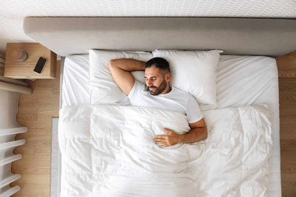 Widok na śpiącego faceta w średnim wieku, cieszącego się spokojną drzemką w łóżku, trzymającego rękę za głowę, we wnętrzu sypialni. Sen i relaks dla koncepcji zdrowia - Zdjęcie, obraz