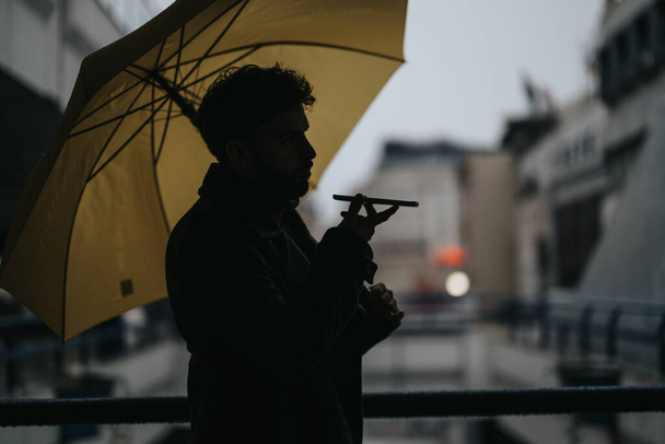 Uomo d'affari che fa una telefonata con il cliente in vivavoce mentre tiene in mano l'ombrello in una giornata piovosa. - Foto, immagini