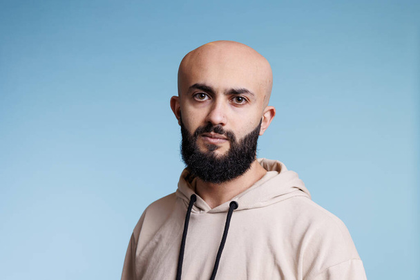 Young bald arab man with relaxed facial expression studio portrait. Bearded arabian person with neutral emotions wearing casual beige hoodie and looking at camera on blue background - Photo, Image