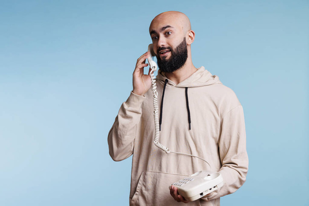 Jeune homme arabe excité tenant un téléphone fixe et communiquant. Personne barbu chauve émotionnelle répondant à un appel téléphonique rétro et parlant avec expression faciale joyeuse - Photo, image