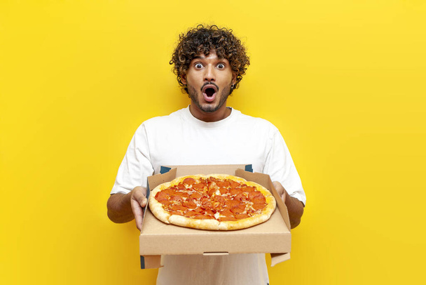 hambriento hindú chico sosteniendo una caja con deliciosa pizza y sorprendido en amarillo aislado fondo, impactado hindú hombre comer y publicidad de comida rápida en asombro - Foto, imagen