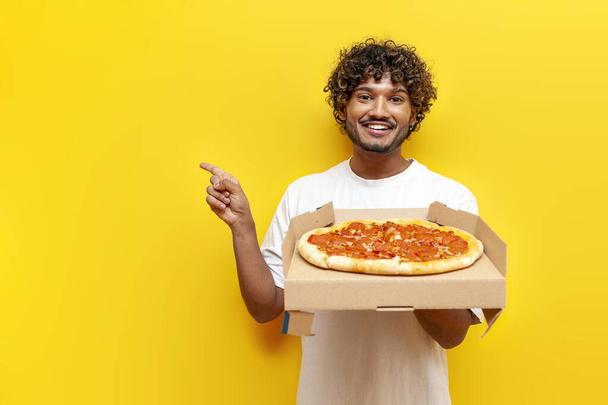 hladový chlap indián drží krabici s lahodnou pizzou a ukazuje s rukou na kopírovat prostor na žlutém izolovaném pozadí, mladý indián jí a inzeruje rychlé občerstvení - Fotografie, Obrázek