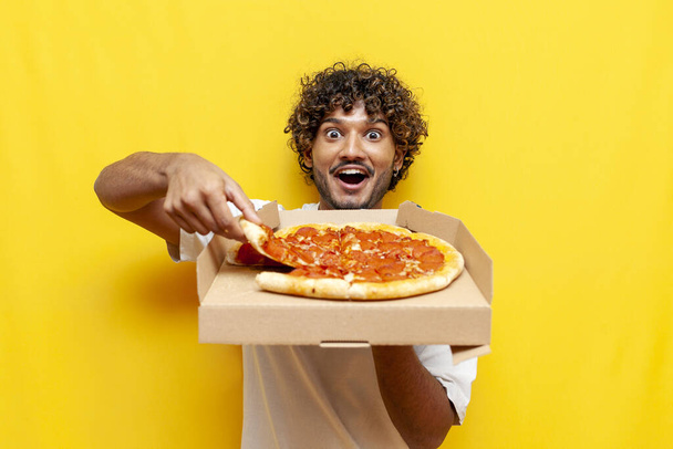 голодний індуський хлопець тримає коробку зі смачною піцою і здивований на жовтому ізольованому фоні, шокований індуїст їсть і рекламує фаст-фуду в подиві - Фото, зображення
