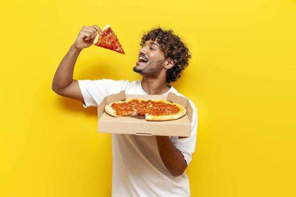 głodny facet indyjski trzyma pudełko z pyszne pizzy i gryzie kawałek na żółtym odizolowanym tle, młody mężczyzna indyjski je i reklamuje fast food - Zdjęcie, obraz