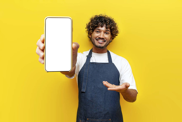 giovane ragazzo cameriere indiano in grembiule mostra schermo bianco di smartphone su sfondo giallo isolato, maschio barista indiano in uniforme pubblicizza cellulare online - Foto, immagini