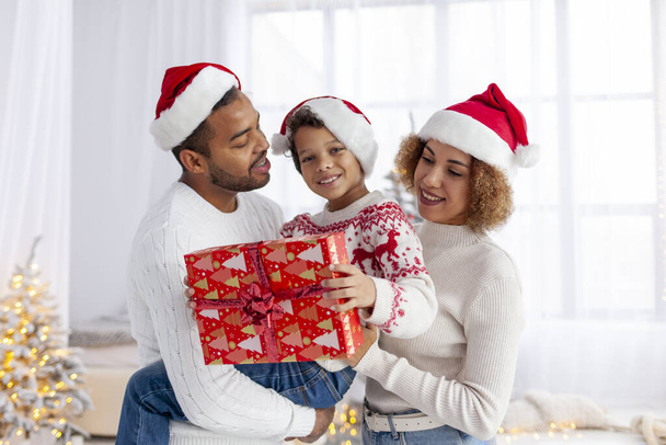 African-American family in Santa hats hugging and smiling at home against the background of christmas tree, teenage boy holds gift box and celebrates the winter holidays with his parents together - Photo, Image