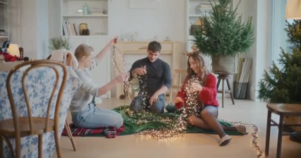 Молоді друзі чоловічої та жіночої статі сортують заплутані освітлені вогні для різдвяних прикрас вдома - Кадри, відео