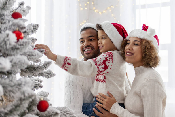 Afro-Amerikaanse familie viert Kerstmis met hun kind en versiert de kerstboom thuis, 10-jarige jongen met zijn ouders in Santa hoed heeft decoratief speelgoed en helpt - Foto, afbeelding