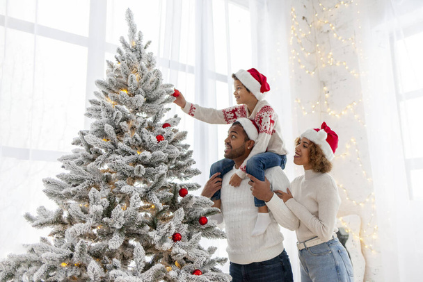 Afro-americká rodina slaví Vánoce se svým dítětem a zdobí vánoční stromeček doma, desetiletý chlapec se svými rodiči v Santa klobouku drží dekorativní hračku a pomáhá - Fotografie, Obrázek