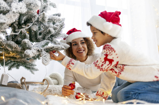 African American teenage boy in Santa hat and Christmas sweater decorates Christmas tree with mom at home, child holds decoration toy and celebrates New Year with parent - Photo, Image