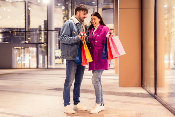 Millennial Ehemann überrascht seine Frau mit einer kleinen verpackten Schachtel und präsentiert sie lächelnd vor dem Einkaufszentrum, in der Hand Einkaufstüten. Romantische Bescherung am Winterabend - Foto, Bild