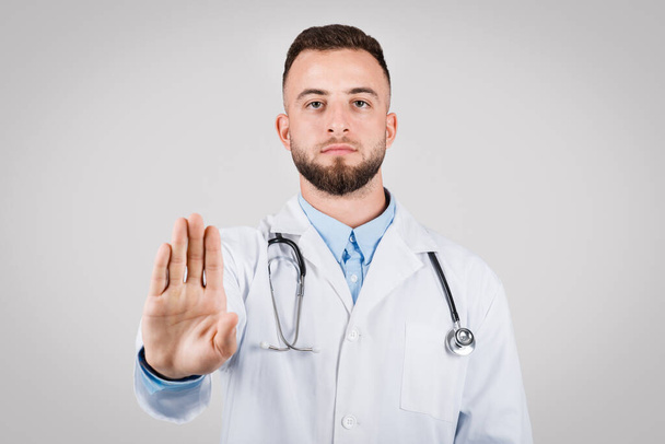 Médico masculino serio con bata de laboratorio que muestra el gesto de stop hand, haciendo hincapié en la importancia de la seguridad sanitaria y el cumplimiento del paciente con el asesoramiento médico - Foto, Imagen