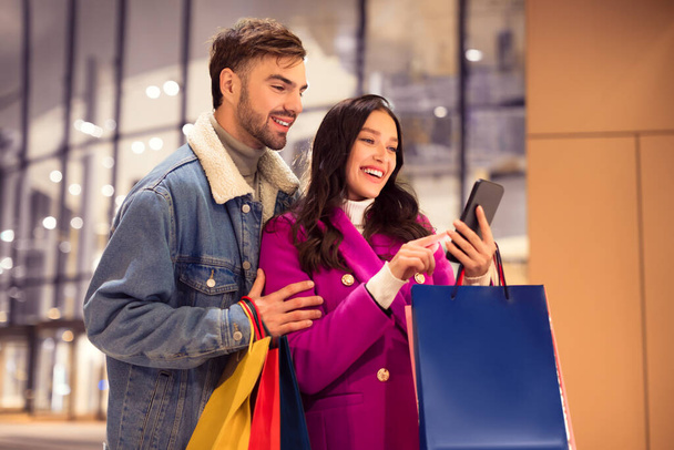 Счастливые молодые клиенты пары со многими сумками смотреть на смартфон, в поисках больших сделок зимний отпуск и продаж, стоя за пределами современного торгового центра вместе. Рождественское веселье - Фото, изображение