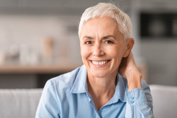 Retrato de mulher madura europeia alegre com cabelo curto cinza sorrindo para a câmera, sentado descansando no sofá em casa interior, vestindo camisa azul estilo casual. Beleza e elegância sem idade - Foto, Imagem