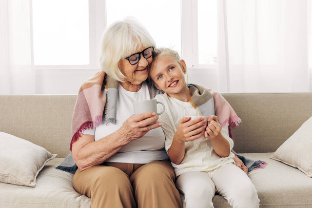 祖母の女性は,家に置かれたシニア娘高齢者が子供のソファー家族を愛し,幸せな抱擁のソファー古い孫娘ハグマグカップライフスタイルガールの冬 - 写真・画像