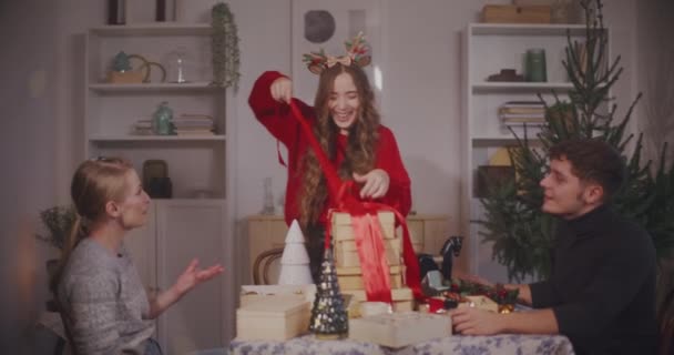 Glückliche junge Frau entfernt Schleife aus Geschenkboxen von männlichen und weiblichen Freunden im Wohnzimmer - Filmmaterial, Video