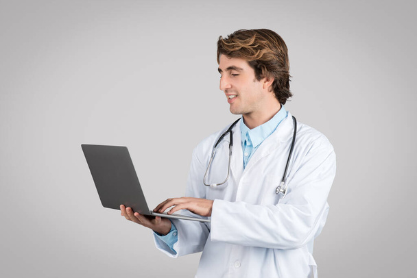 Confident male doctor in white coat with stethoscope typing on laptop isolated on gray background, studio. Online consultation, medical service remote, health care - Photo, Image