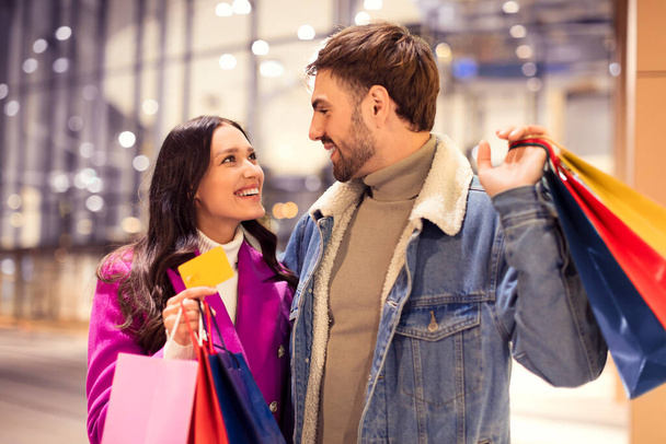 Lady zeigt dem Mann die Kreditkarte, wenn er mit dem Wintereinkauf beginnt, und umarmt ihn nachts vor dem Einkaufszentrum. Paar in Winterjacken findet die perfekten Geschenke für Weihnachten und Valentinstag - Foto, Bild
