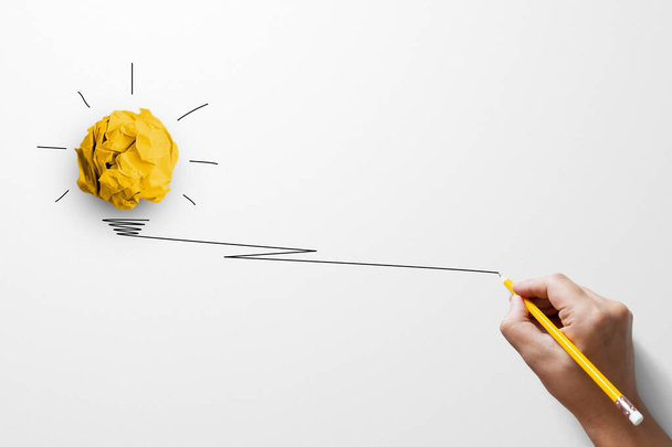 Δημιουργικές ιδέες σκέψης και έννοια της καινοτομίας. Σφαίρα απορριμμάτων χαρτιού κίτρινο χρώμα με το σύμβολο του λαμπτήρα στο λευκό φόντο και το χέρι κρατώντας κίτρινο μολύβι - Φωτογραφία, εικόνα