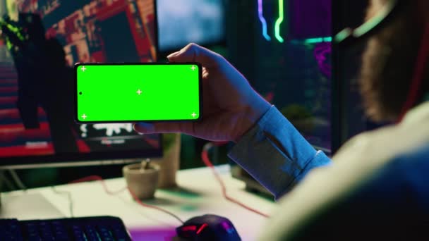Man in dimly lit apartment watching tutorial on how to finish videogame on green screen smartphone, enjoying day off. Gamer learning online multiplayer shooter tips from video on mockup mobile phone - Footage, Video