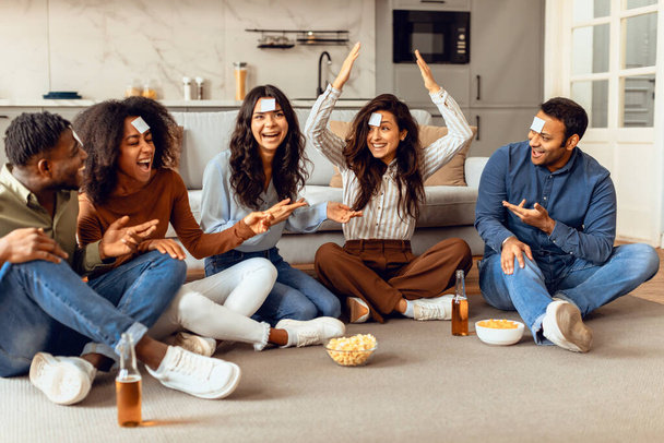 Fröhliche multikulturelle Studentenfreunde beim Ratespiel, genießen Bier und Popcorn, sitzen auf dem Fußboden im modernen Wohnzimmer, chillen am Wochenende zu Hause zusammen, haben Karten auf der Stirn - Foto, Bild