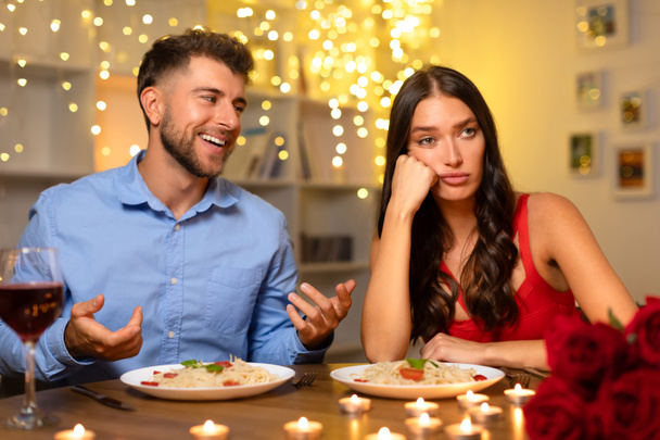 Beim Abendessen bei Kerzenschein spricht der fröhliche Mann lebhaft, während die Frau im roten Kleid unbeeindruckt erscheint, ihre Wange auf die Hand legt und am Esstisch sitzt - Foto, Bild