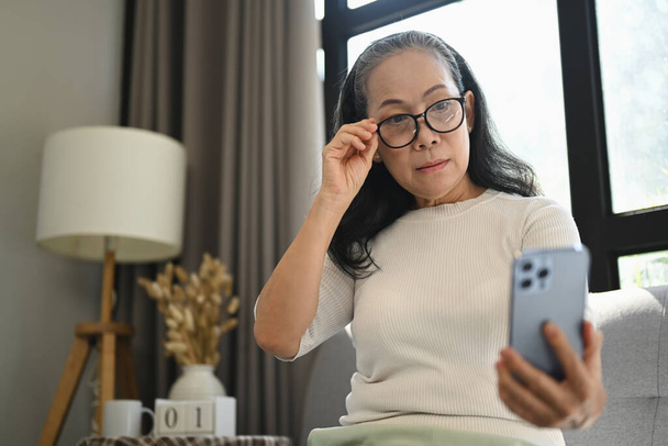 Beautiful middle age woman in glasses checking apps on mobile phone while sitting on couch. - 写真・画像