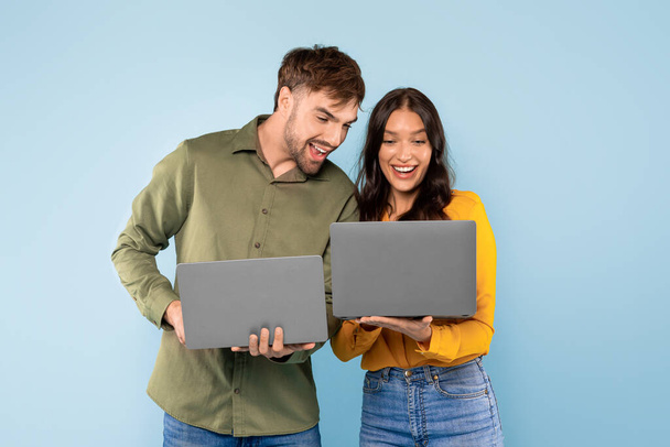 Jovem casal alegre comparando notas em seus laptops, compartilhando uma risada sobre uma descoberta digital contra um fundo azul brilhante, navegar na internet - Foto, Imagem