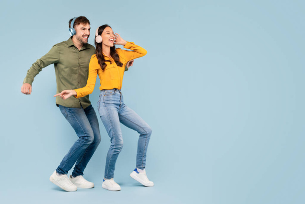 Joven alegre hombre y mujer en ropa casual con auriculares disfrutar de la música mientras bailan juntos contra un fondo azul sólido, espacio libre - Foto, imagen