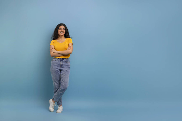 Cute pozytywne dość kręcone młoda Hinduska kobieta w casual ubrania trzymając się za ręce skrzyżowane na klatce piersiowej, patrząc na przestrzeń ksero dla reklamy i uśmiechnięty, niebieskie tło studio, pełna długość - Zdjęcie, obraz