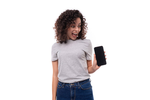 portrait of a young surprised joyful 30 year old caucasian woman in a gray t-shirt holding a smartphone with a mockup on a white background with copy space. - Photo, Image