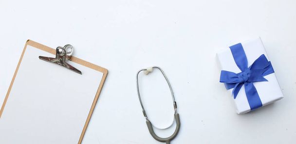 Κάτοψη και επίπεδη θέσει, αντιγραφή χώρο γραφείο γιατρούς γραφείο. Ιατρικά εξαρτήματα, Λευκό ιατρικό πρόχειρο, στηθοσκόπιο και χάρτινο κουτί δώρου χειροτεχνίας με μπλε κορδέλα φιόγκο, γυαλιά, απομόνωση στυλό σε λευκό φόντο - Φωτογραφία, εικόνα