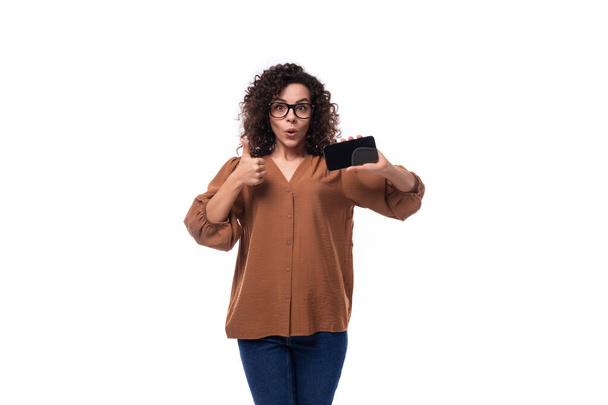 junge europäische lockige Frau mit schwarzen Haaren in einer Bluse zeigt den Bildschirm eines Smartphones mit einer Attrappe. - Foto, Bild