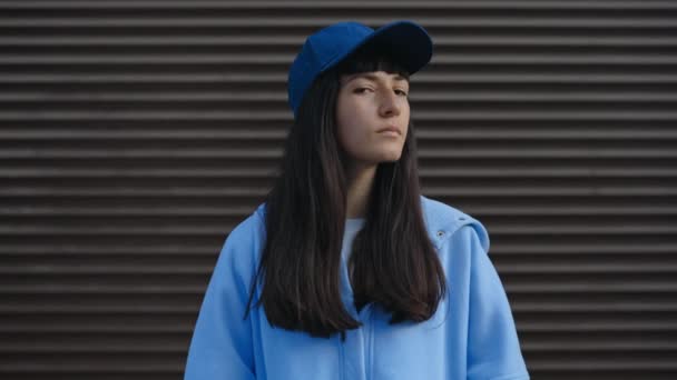 Una joven seria con el pelo castaño, con una gorra azul y ropa casual, se encuentra en una calle de la ciudad. Este retrato al aire libre la captura como turista o viajera en un entorno urbano - Imágenes, Vídeo