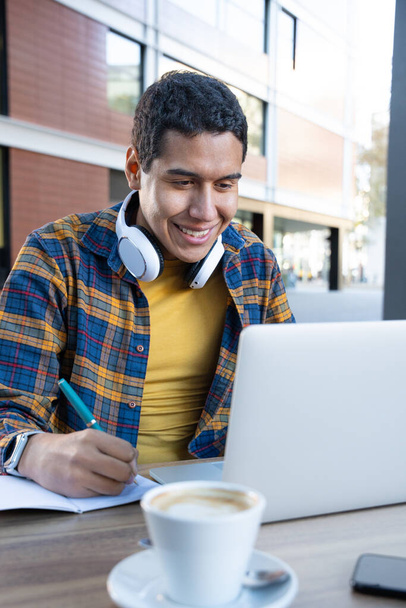 Függőleges fotó egy fiatal latin férfiról, aki egy szabadtéri büfében tanul reggel, jegyzetfüzetre ír és laptopot használ egy csésze kávéval a táblában. - Fotó, kép