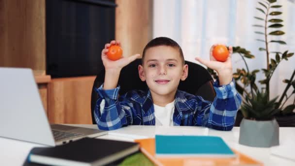 Grundschüler zeigen Daumen hoch mit Mandarinen in den Händen, während sie am Schreibtisch mit Laptop und bunten Notizbüchern sitzen. Lächelndes Kind im karierten Hemd macht Pause für Snacks zwischen Online-Unterricht. - Filmmaterial, Video