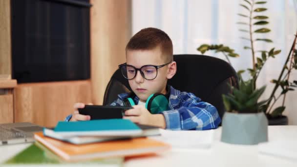 Przystojny biały dzieciak siedzi przy stole z nowoczesnym smartfonem i bezprzewodowymi słuchawkami podczas przerwy w pracy domowej. Poważny mężczyzna dziecko odpoczywając po odległość uczenie się w pomieszczeniach. - Materiał filmowy, wideo