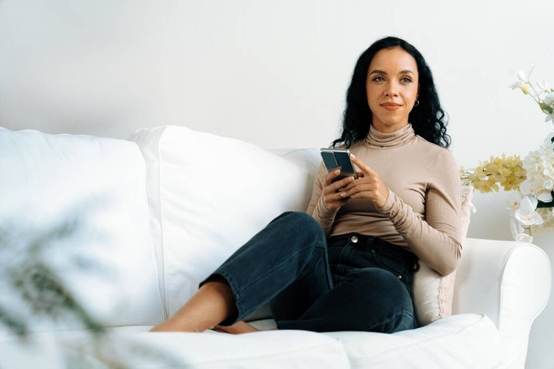 Uvolněná mladá Afroameričanka pomocí klíčového mobilního telefonu na pohovce v obývacím pokoji doma - Fotografie, Obrázek