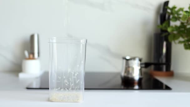 Vor dem Hintergrund einer modernen Küche wird weißer Rohreis in Zeitlupe in ein transparentes Gefäß gegossen. Gesunde Ernährung und Ernährungskonzept. Sichere, ökologische und gesunde Produkte. Ökologisch - Filmmaterial, Video