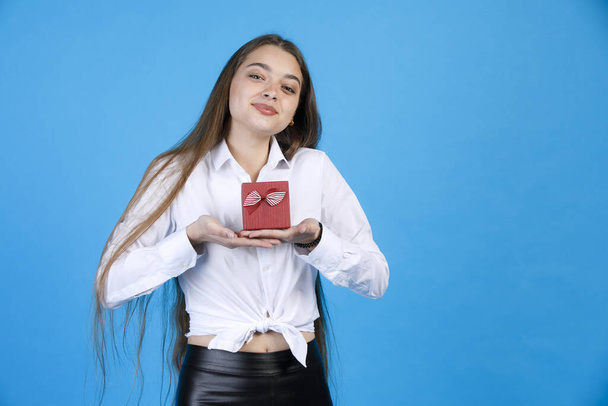 Cheerful pretty girl sincerely looking at camera, while raising tiny gift box with both hands. Portrait of happy woman in white blouse demonstrating present, isolated on blue. Concept of birthday. - Photo, Image