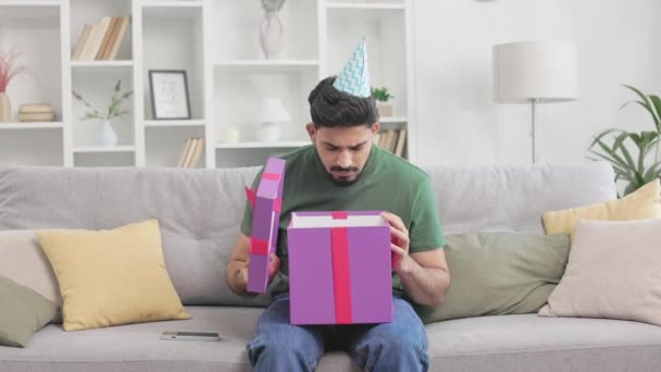 Enttäuschter indischer Mann öffnet verpackte Geschenkschachtel mit roter Schleife und sieht darin etwas Unangenehmes. Trauriger Kerl nimmt Partyhut ab und ist traurig über festliche Überraschung. - Filmmaterial, Video