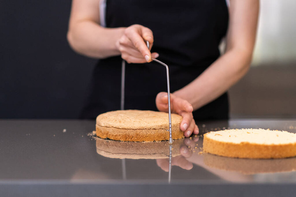 Femme qualifiée utilisant un couteau à ficelle pour séparer les couches de gâteau dans la cuisine moderne confiseur femelle en tablier noir debout à grande table faisant des confiseries personnalisées - Photo, image