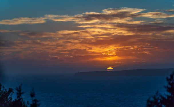 Ein Sonnenuntergang erhellt ein endloses Meer, wie ein Leuchtturm auf Klippen steht. - Foto, Bild