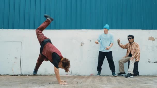 Junge asiatische Tänzerin tanzt im Hip-Hop-Stil, während multikulturelle Freunde hinter der Mauer jubeln. Attraktiver gutaussehender Mann streckt die Arme auf der Straße mit blauem Hintergrund aus. Outdoor-Sport 2024. hiphop. - Filmmaterial, Video