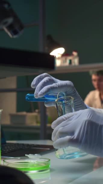 Tanımlanamayan kadın bilim adamının ellerinin dikey görüntüsü mavi sıvıyı mataraya döküyor, yeşil substrat ekliyor, sonra içerikleri karıştırıyor ve sonucu kontrol ediyor. - Video, Çekim