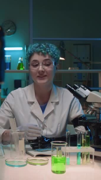 Közepes függőleges lövés fiatal női tudós fehér köpenyben, szemüveg, műkar ül a kémiai laborban, mutatja lombik és üveg hajlító oldat, miközben rögzíti blog tartalmát a kamera - Felvétel, videó