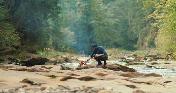 Ένας άντρας στο δάσος κοντά σε μια φωτιά αναπαύεται κατά τη διάρκεια ενός τουριστικού ταξιδιού. Πεζοπορία στο δάσος ή στα βουνά ενός μόνο ταξιδιώτη. Ένας τουρίστας σε ένα τουριστικό στρατόπεδο - Φωτογραφία, εικόνα
