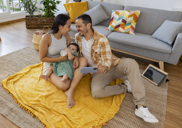 Pohled shora z usměvavé latinské rodiny sedí na podlaze svého obývacího pokoje přičemž selfie - Fotografie, Obrázek